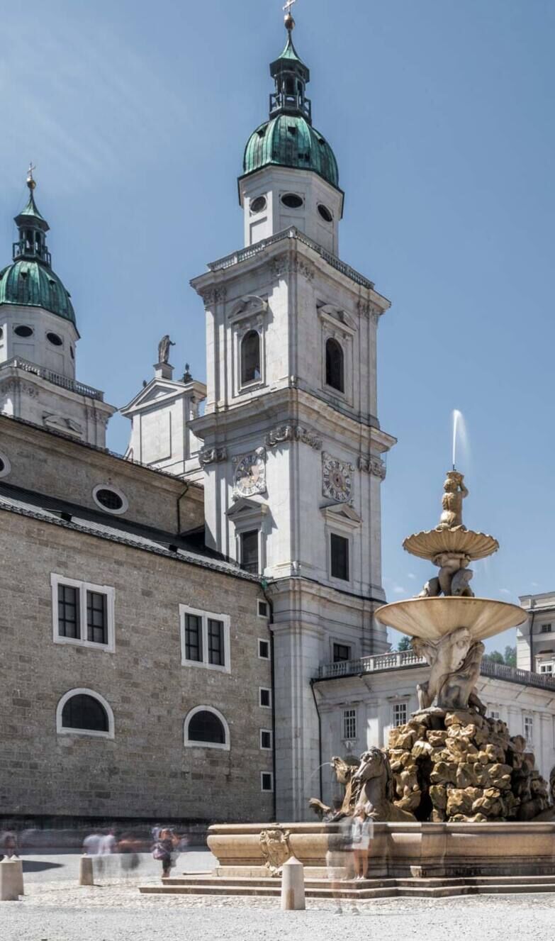 Salzburger Domplatz mit historischen Gebäuden und einem imposanten Brunnen unter blauem Himmel.