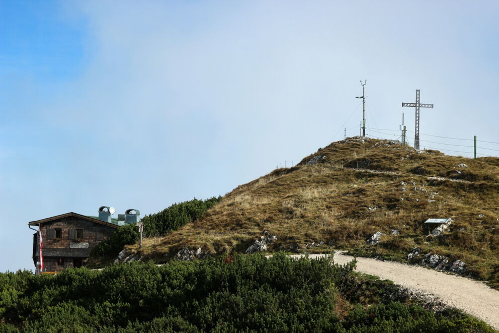 Almhütte neben einem Gipfelkreuz auf einem Hügel unter blauem Himmel.