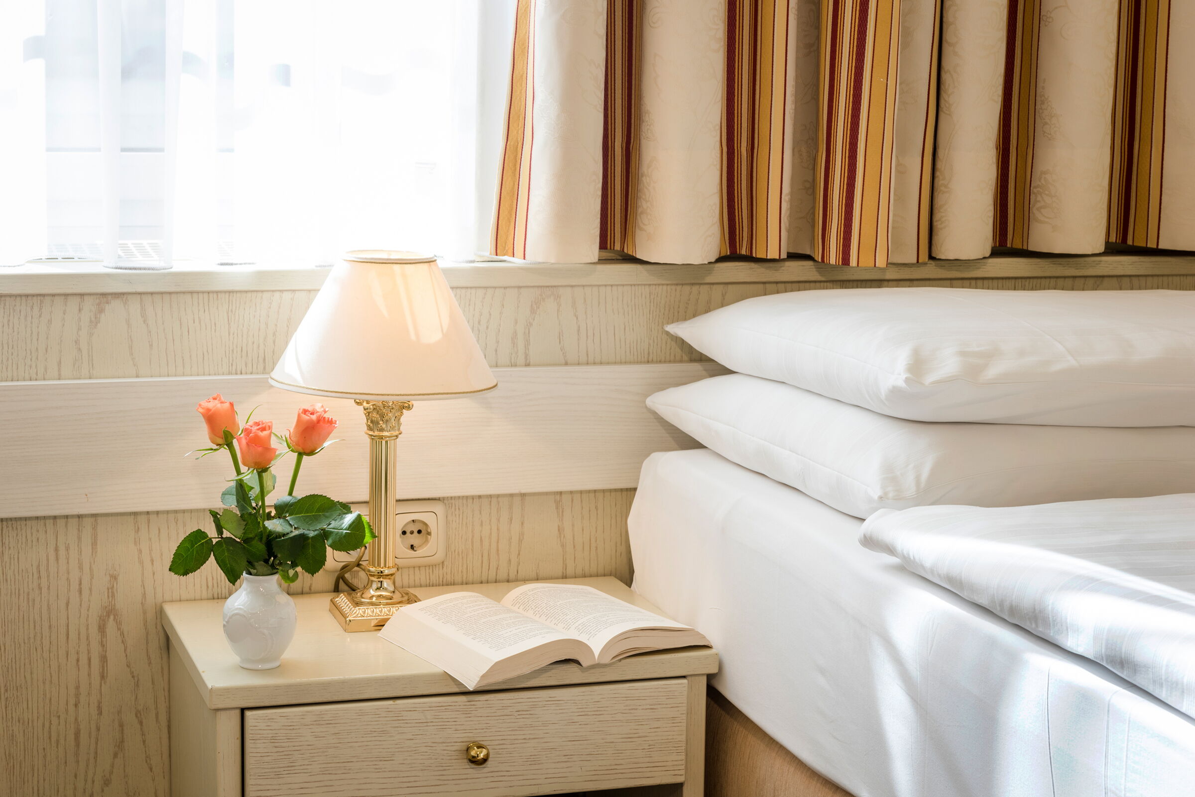 Helles Zimmer mit Bett, Nachttisch, Lampe, Buch und Vase mit Rosen.