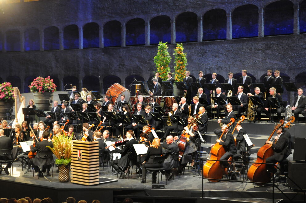 Orchestra plays in a historical setting, musicians in elegant formation.
