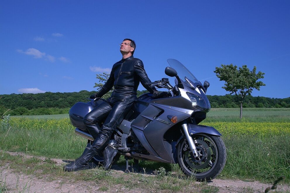 Mann in schwarzer Lederkleidung sitzt auf Motorrad vor grünem Feld und blauem Himmel.