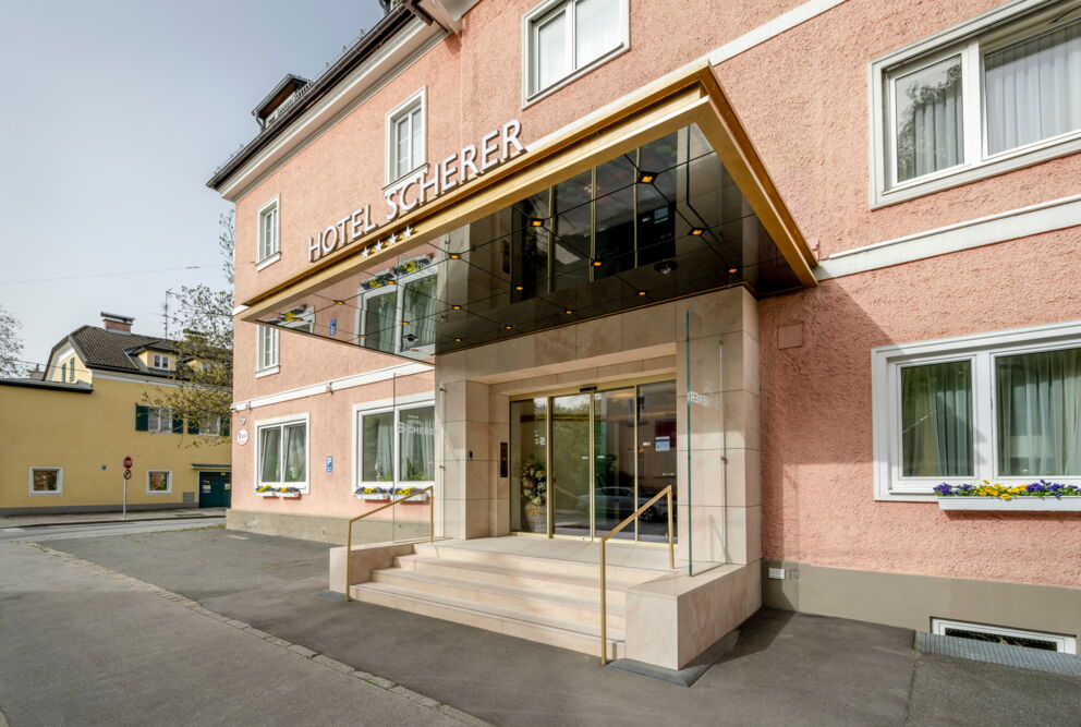 Eingang des Hotels Scherer mit Glasüberdachung und blühenden Blumenkästen.