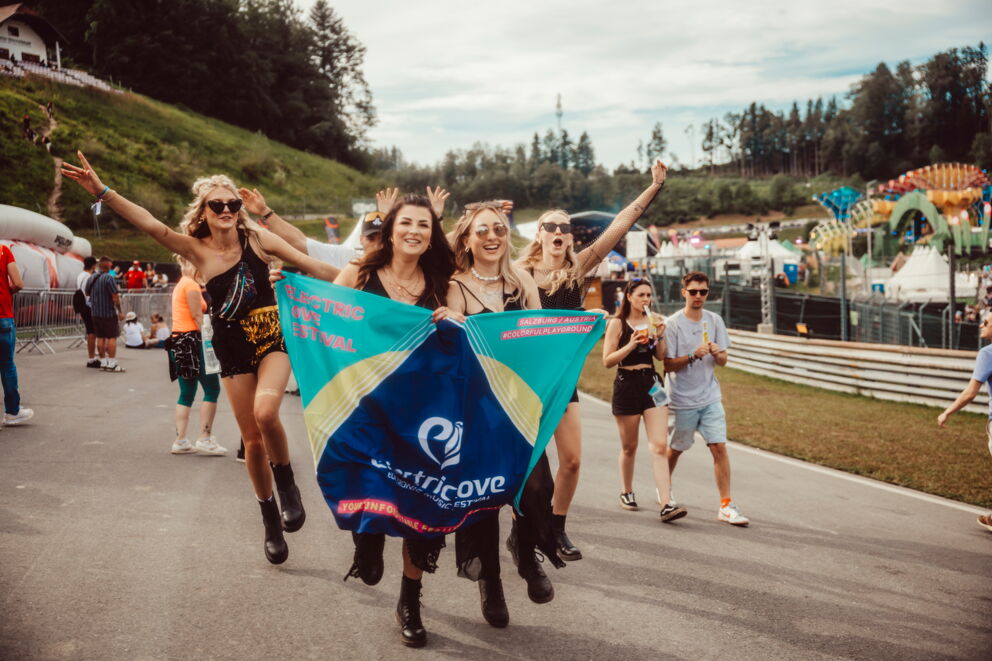 Menschen tanzen fröhlich mit Festival-Flagge in sonniger Umgebung.