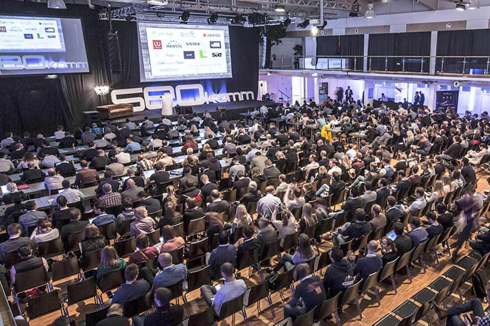 Large crowd in a conference hall with presentation on stage.