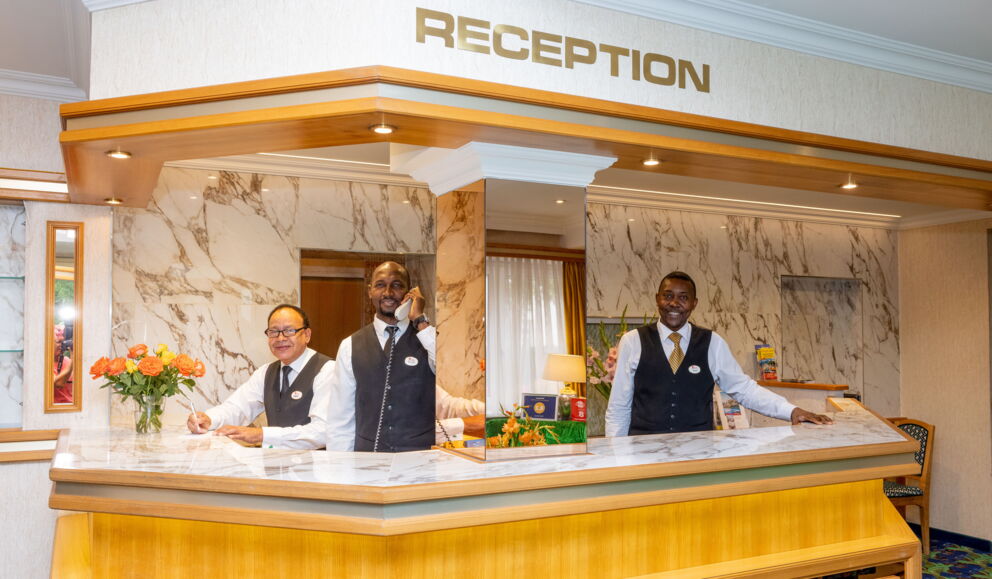 Drei Hotelangestellte an der Rezeption mit Marmorverkleidung und Blumenarrangement.