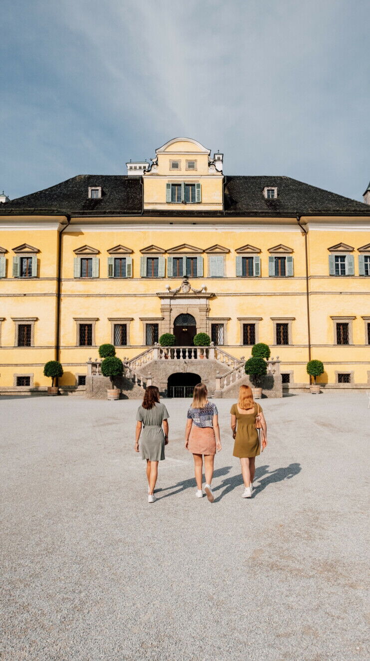 Three people are walking towards an impressive yellow castle with stone steps.