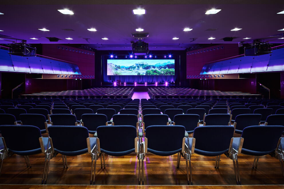 Großer, beleuchteter Konferenzraum mit Sitzreihen und Präsentationsleinwand in einem Hotel.