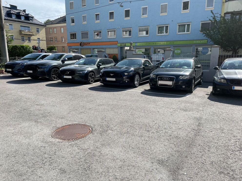Mehrere Autos parken nebeneinander auf einem sonnigen Parkplatz vor einem blauen Gebäude.