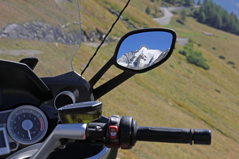 Motorcycle handlebars with mirror view of snow-covered mountain and winding road.