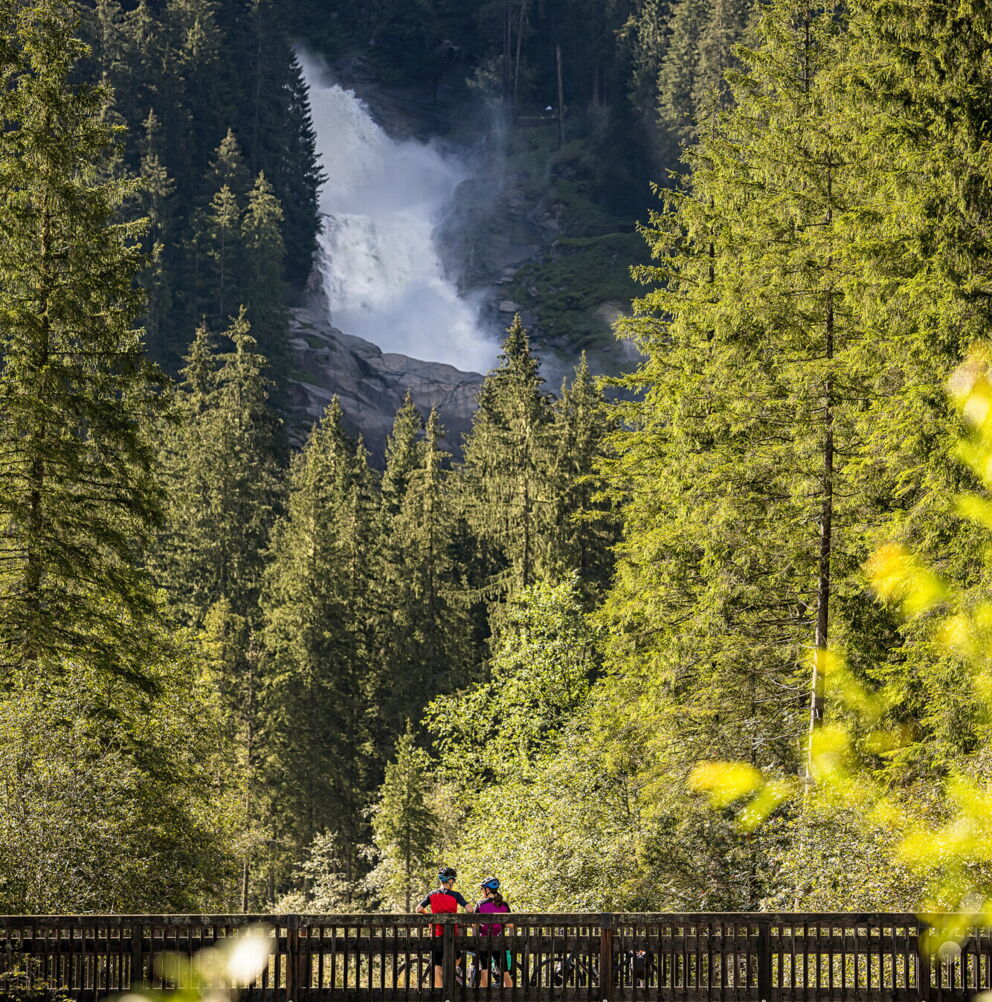 Zwei Radfahrer auf Holzbrücke mit Wasserfall im Hintergrund, umgeben von dichten Wäldern.