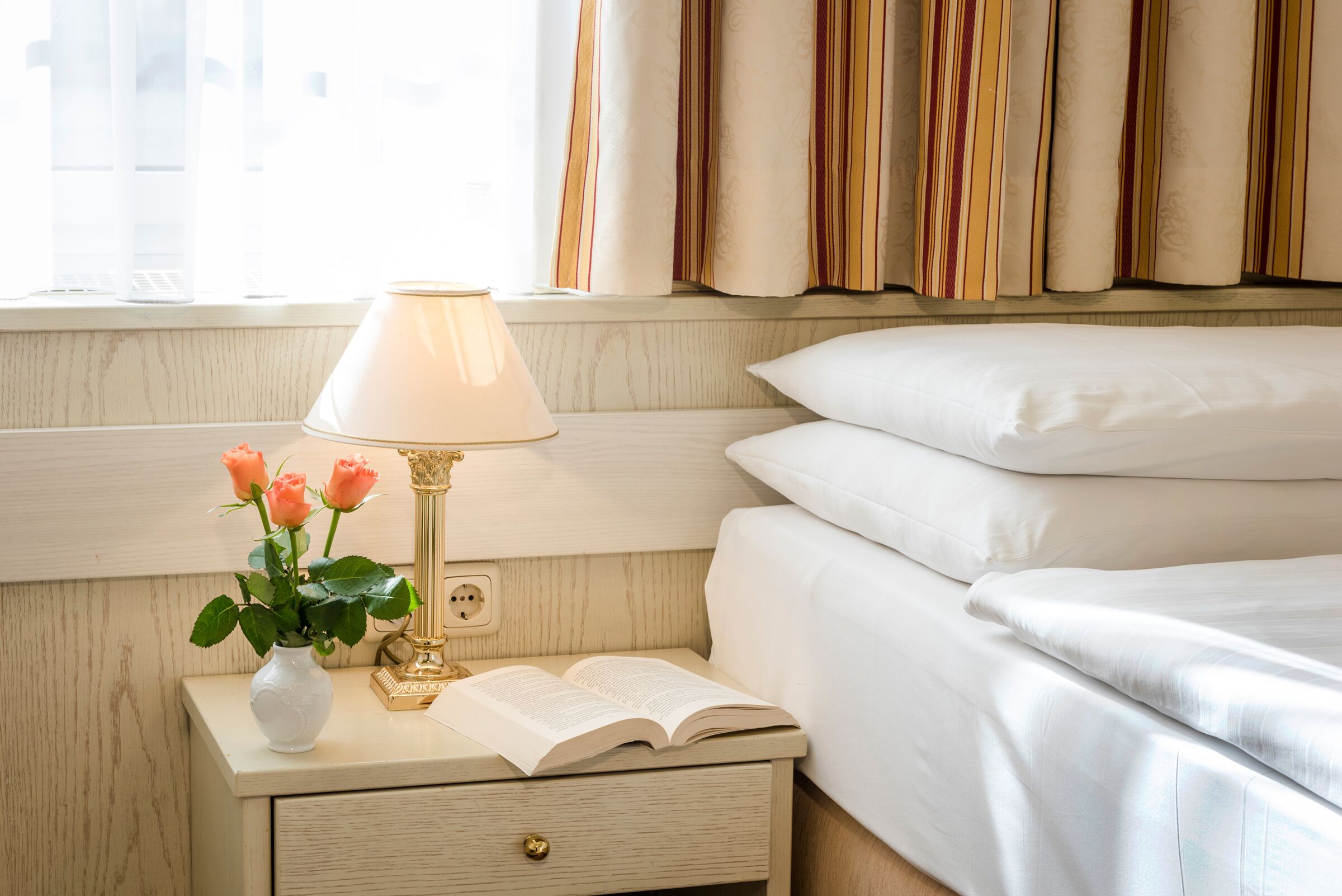 Helles Zimmer mit Bett, Nachttisch, Lampe, Buch und Vase mit Rosen.