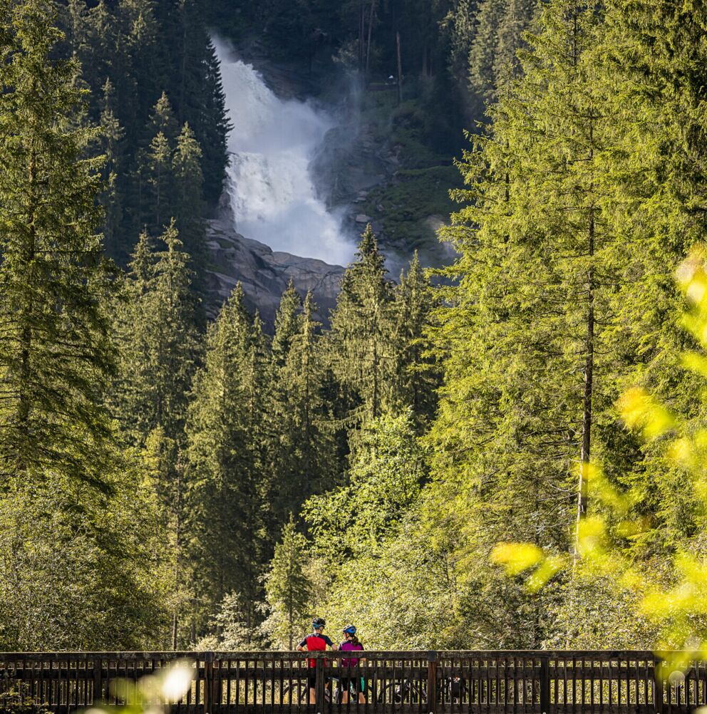 Zwei Radfahrer auf Holzbrücke mit Wasserfall im Hintergrund, umgeben von dichten Wäldern.
