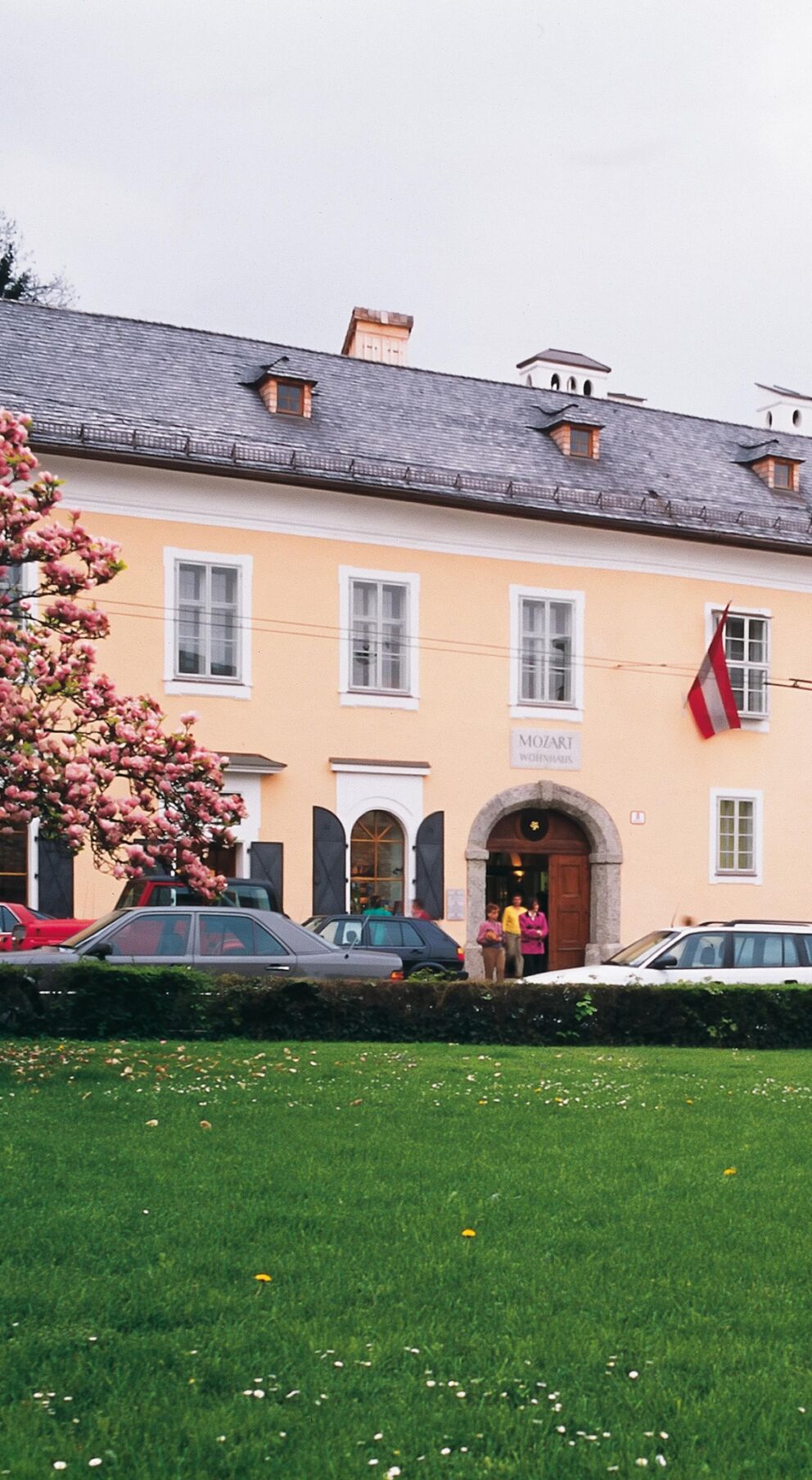 Historisches Gebäude mit gepflegtem Garten, blühenden Bäumen und parkenden Autos davor.