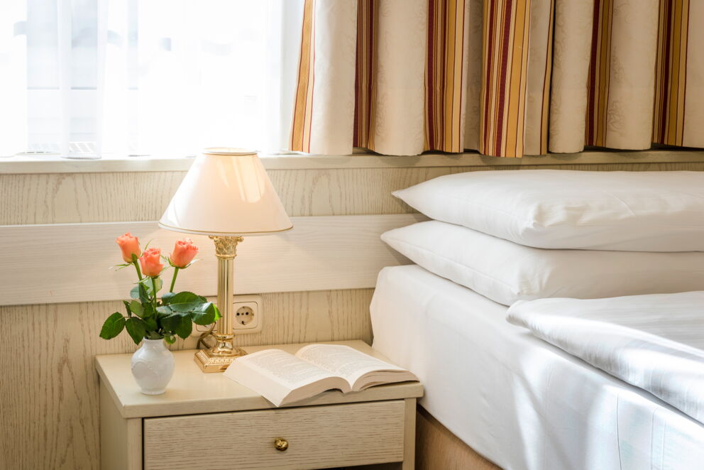 Helles Zimmer mit Bett, Nachttisch, Lampe, Buch und Vase mit Rosen.