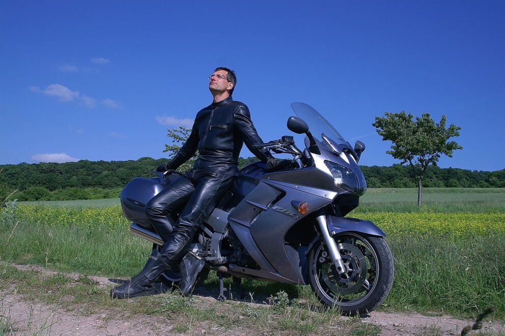 Man in black leather clothing sits on a motorcycle in front of a green field and blue sky.