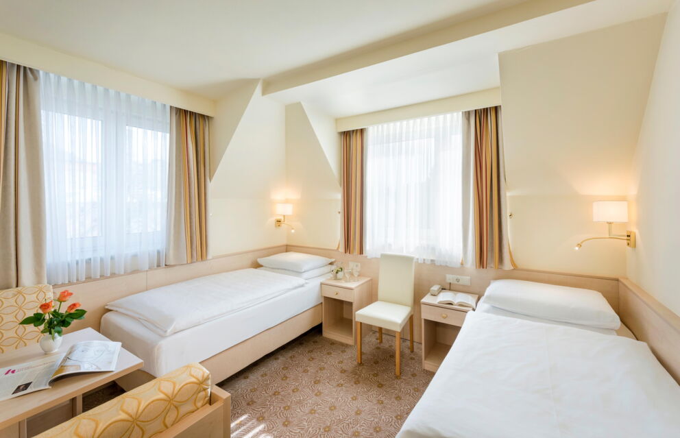 Helles Zimmer mit zwei Einzelbetten, Tisch, Stühlen, Fenstern, Vorhängen und Blumendekoration.