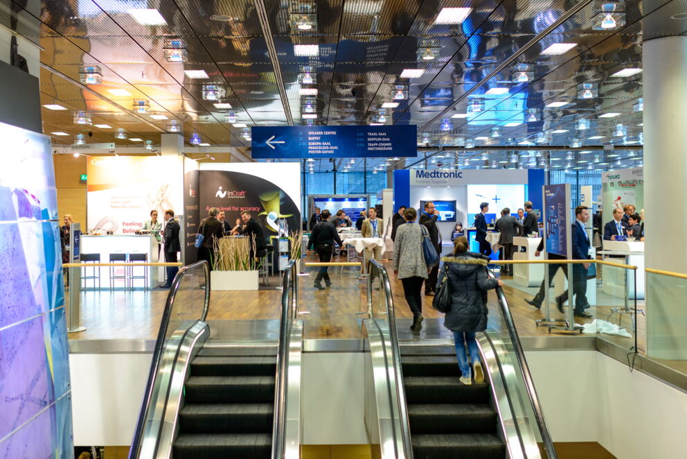 Menschen auf einer Messe mit Ständen und Rolltreppe im Vordergrund.