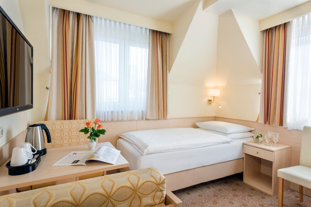 Helles Einzelzimmer mit gemütlichem Bett, frischen Blumen und stilvollen Vorhängen.