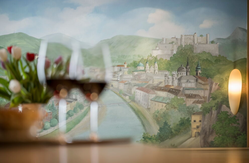 Zwei Weingläser vor einem malerischen Wandbild der Stadt Salzburg.