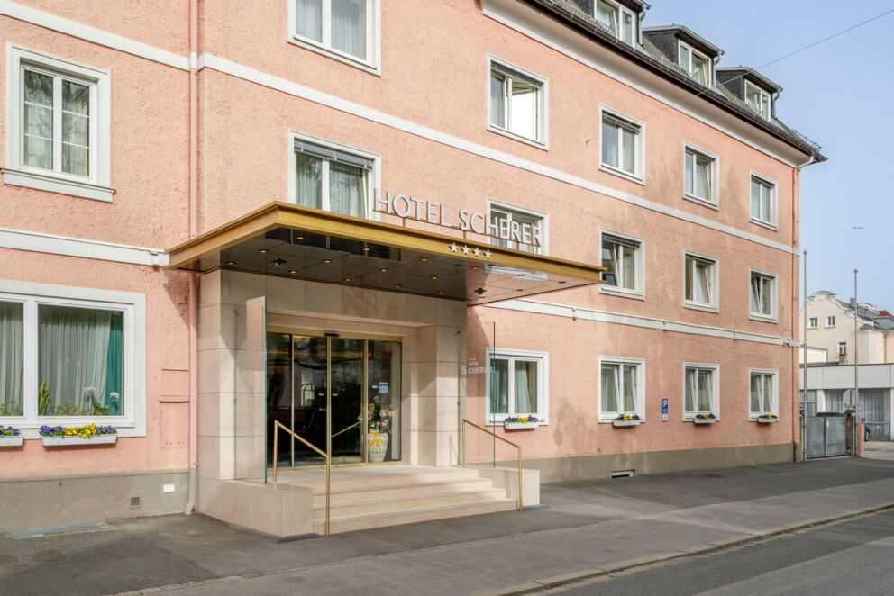 Hoteleingang mit goldener Überdachung, großen Fenstern und blühenden Blumenkästen.