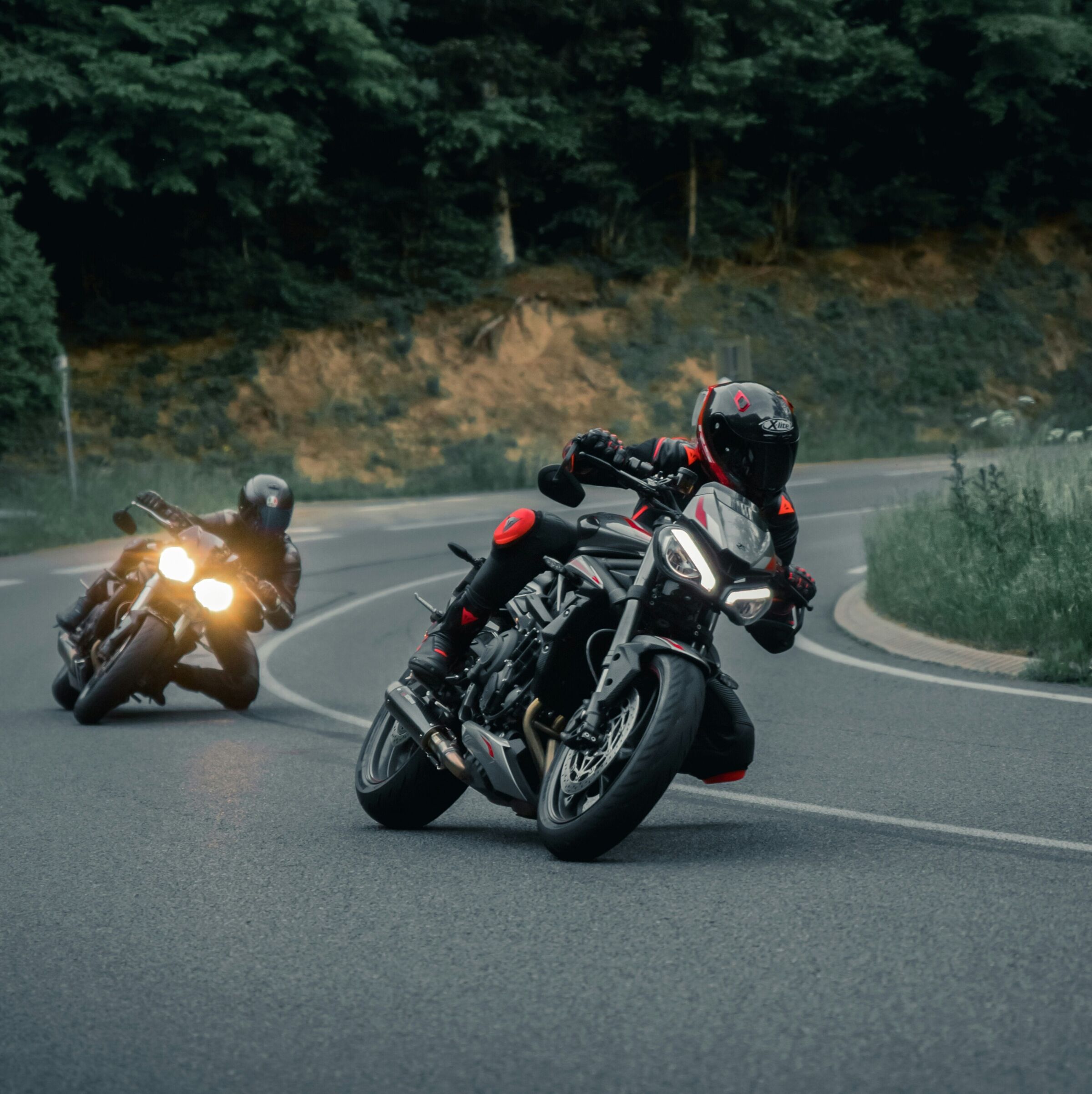 Zwei Motorradfahrer in Kurve auf offener Straße umgeben von Bäumen.