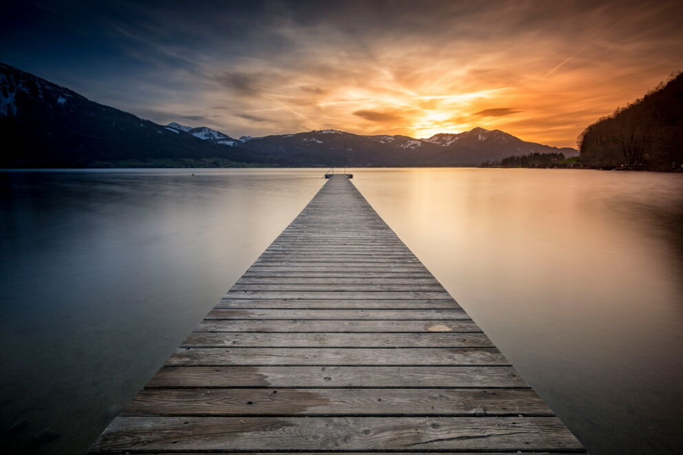 Wooden footbridge leads into a calm lake with a spectacular sunset and mountain backdrop.