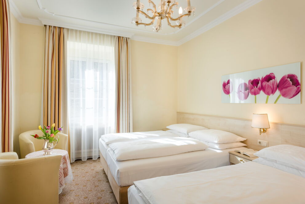 Helles Zimmer mit zwei Betten, Blumenbild, Kronleuchter und gemütlicher Sitzgelegenheit.