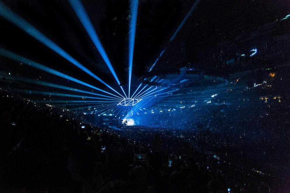 Bright lights and an enthusiastic crowd at a concert in a large hall.
