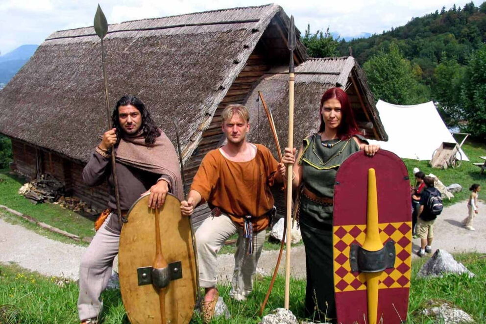 Three people in historical costumes in front of traditional huts in a green landscape.